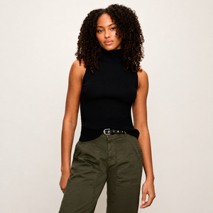 A woman with curly dark hair wearing a black sleeveless mock neck top and olive green cargo pants, standing against a light gray studio background.