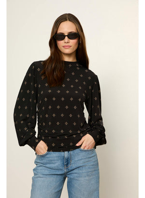A woman modeling a black long-sleeve blouse with a small beige geometric pattern, paired with light blue jeans and black sunglasses, against a plain light beige background.