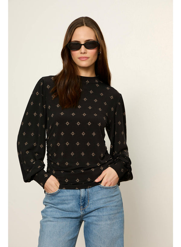 A woman modeling a black long-sleeve blouse with a small beige geometric pattern, paired with light blue jeans and black sunglasses, against a plain light beige background.