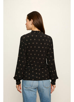 A woman viewed from the back wearing a black long-sleeve blouse with a small beige geometric pattern and light blue jeans, standing against a plain off-white background.