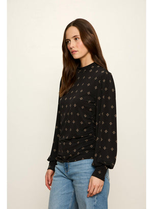 A woman wearing a black long-sleeve top with a small beige geometric pattern and light blue jeans, photographed against a plain off-white studio background.