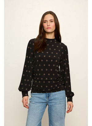 A woman modeling a black long-sleeve top with a small beige geometric pattern, paired with light blue jeans, against a plain light beige background.