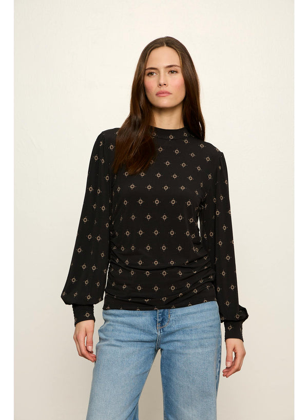 A woman modeling a black long-sleeve top with a small beige geometric pattern, paired with light blue jeans, against a plain light beige background.