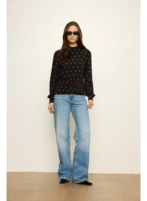 A woman modeling a black long-sleeve top with a small gold geometric pattern, paired with light-wash blue jeans and black flats, standing against a plain off-white wall.