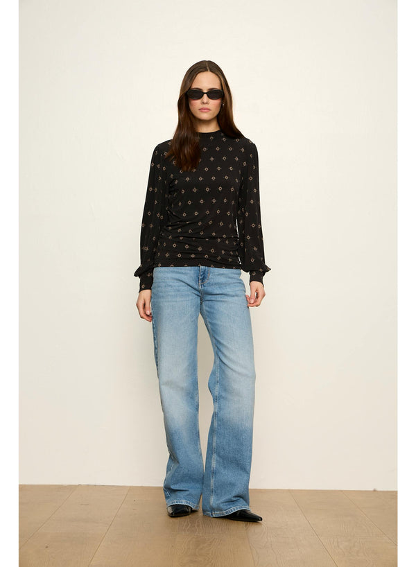 A woman modeling a black long-sleeve top with a small gold geometric pattern, paired with light-wash blue jeans and black flats, standing against a plain off-white wall.