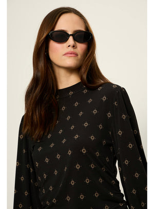 A woman with long brown hair wearing black oval sunglasses and a black long-sleeve top with a small tan geometric pattern, photographed against a plain light beige background.