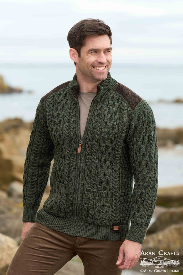 A smiling man wearing a dark olive green cable-knit zip-up sweater with brown shoulder panels and a brown undershirt, standing outdoors near the sea and rocks.