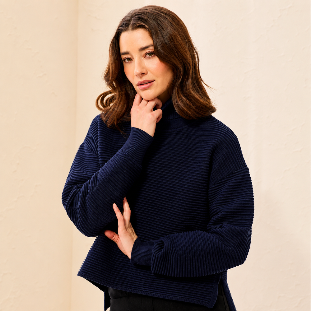 A woman wearing a dark navy ribbed knit mock neck sweater, posing against a textured beige wall.