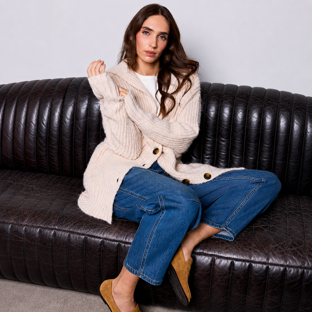 A woman sits on a dark brown leather sofa wearing a chunky cream cable-knit cardigan over a white top, blue jeans, and tan slip-on shoes.