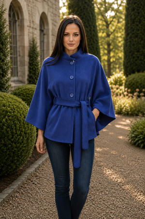 A woman wearing a royal blue cape-style coat with a matching tie belt and dark blue jeans, standing in a garden path with trimmed hedges and a stone building in the background.