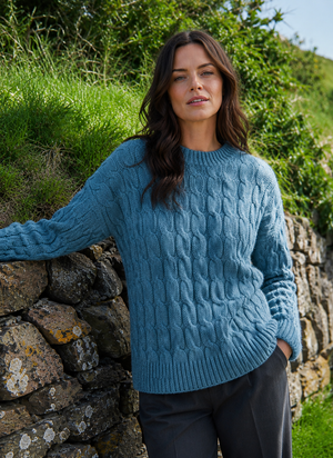 A woman wearing a blue cable-knit sweater and dark trousers, leaning against a stone wall outdoors with green grass and shrubs in the background.