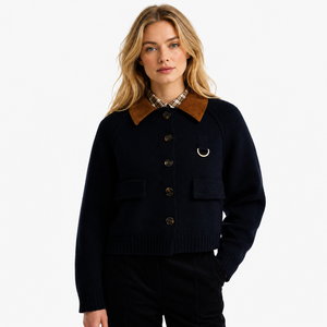 A woman modeling a dark navy knit cardigan with a contrasting brown collar, dark buttons, and a small gold-tone ring detail on the chest, paired with black trousers against a plain white studio background.