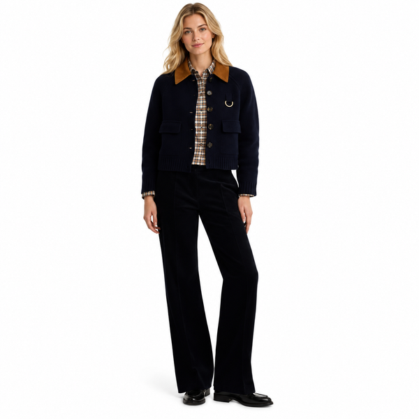 A woman modeling a dark navy knit jacket with brown corduroy collar and a gold ring detail, paired with black trousers and a plaid shirt, on a white studio background.