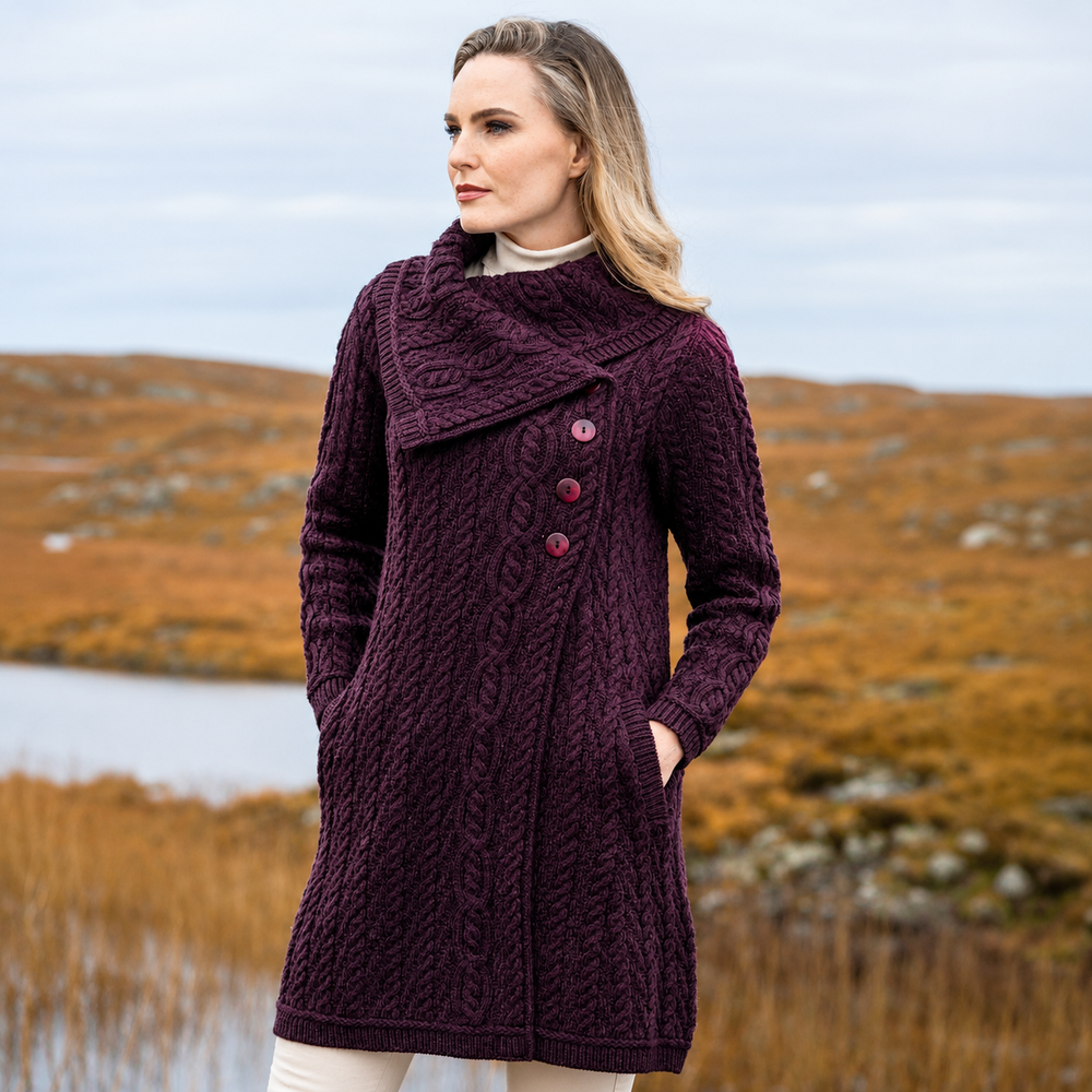 A woman wearing a dark purple cable-knit wrap coat with three buttons, standing outdoors in a grassy, hilly landscape.