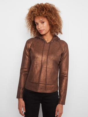 A woman with curly light brown hair wearing a metallic bronze-brown cropped hoodie with a textured, quilted pattern and black pants, standing against a plain light gray background.