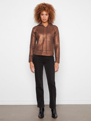 A model with curly light brown hair stands against a white wall wearing a metallic bronze cropped hooded jacket, black pants, and black ankle boots.