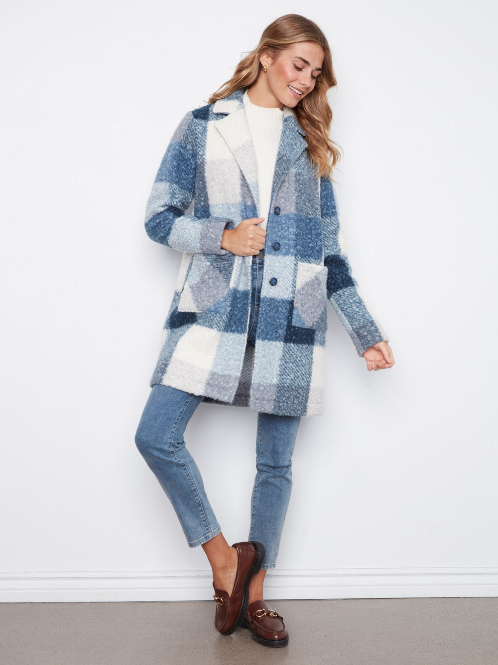 A woman models a blue and white plaid fuzzy cardigan coat over a white top, blue jeans, and brown loafers, standing against a white wall.