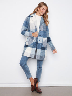 A woman models a blue and white plaid fuzzy cardigan coat over a white top, blue jeans, and brown loafers, standing against a white wall.
