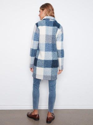 A woman shown from the back wearing a blue and white plaid fuzzy coat, blue jeans, and brown loafers, standing against a plain white wall.
