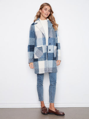 A woman modeling a blue and white patchwork sherpa coat, paired with light blue jeans, a white top, and brown leather loafers, standing against a white wall.