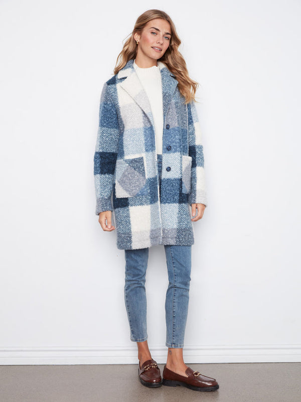 A woman modeling a blue and white patchwork sherpa coat, paired with light blue jeans, a white top, and brown leather loafers, standing against a white wall.