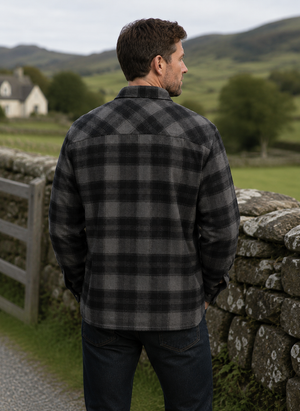 A man viewed from behind wearing a dark gray and black plaid flannel shirt, standing outdoors next to a stone wall with a green rural landscape in the background.