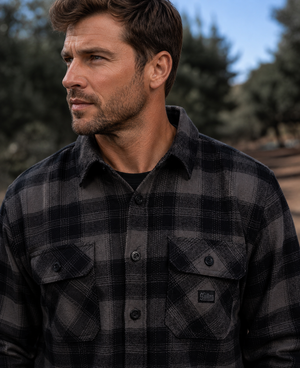 A man wearing a dark gray and black plaid button-down flannel shirt, photographed outdoors with a blurred natural background.