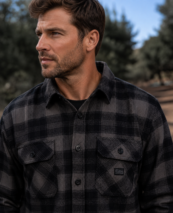 A man wearing a dark gray and black plaid button-down flannel shirt, photographed outdoors with a blurred natural background.