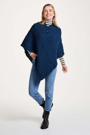 A woman modeling a chunky navy blue cable-knit poncho sweater over a striped turtleneck, blue jeans, and black ankle boots, photographed against a plain white studio background.