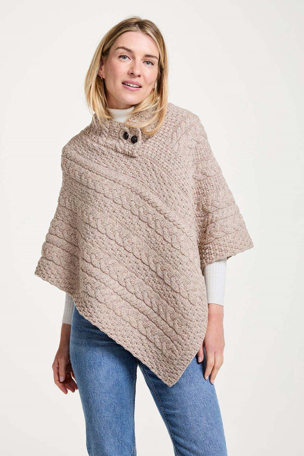 A woman wearing a chunky cable-knit poncho in a light taupe-beige color, styled over a white turtleneck and blue jeans, against a plain off-white studio background.