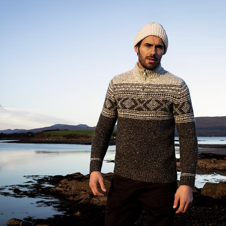A man wearing a cream and dark gray patterned knit sweater, a cream beanie, and dark trousers stands on a rocky shoreline with water and hills in the background.