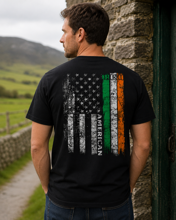 A man wearing a black t-shirt with a distressed graphic of the American flag on the back, featuring vertical stripes in green, white, and orange with the word 'AMERICAN' printed on them, standing outdoors next to a stone wall.