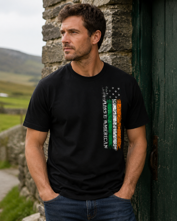 A man wearing a black graphic t-shirt with a vertical distressed flag design in green, white, and orange, and the text 'ANIME AMERICAN' printed alongside it. He is leaning against a rustic stone wall next to a dark green wooden door, with a blurred outdoor landscape in the background.