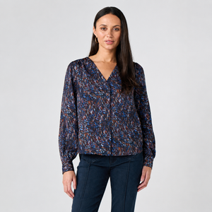 A woman modeling a long-sleeve V-neck blouse with a dense abstract multicolor print in blue, orange, and brown on a dark base, paired with dark blue jeans, against a light gray studio background.