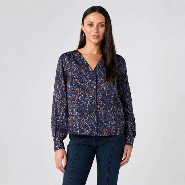 A woman modeling a long-sleeve V-neck blouse with a dense abstract multicolor print in blue, orange, and brown on a dark base, paired with dark blue jeans, against a light gray studio background.