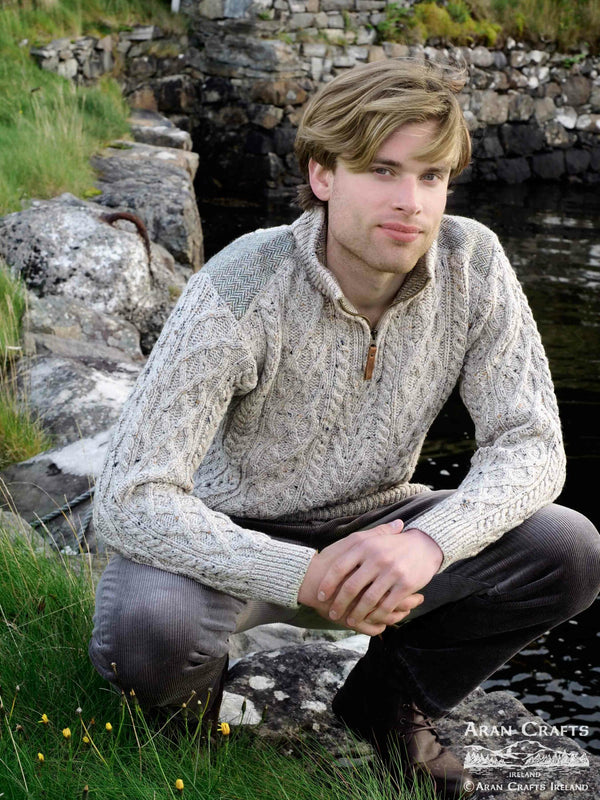 Man wearing a knitted sweater sitting by a stone wall and water.