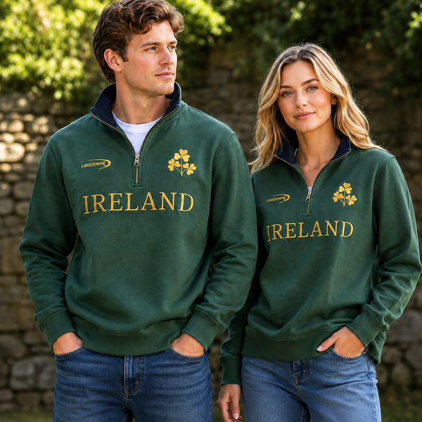 A man and a woman wearing matching dark green quarter-zip pullovers with 'IRELAND' in gold lettering and a shamrock graphic on the chest, standing outdoors in front of a stone wall.