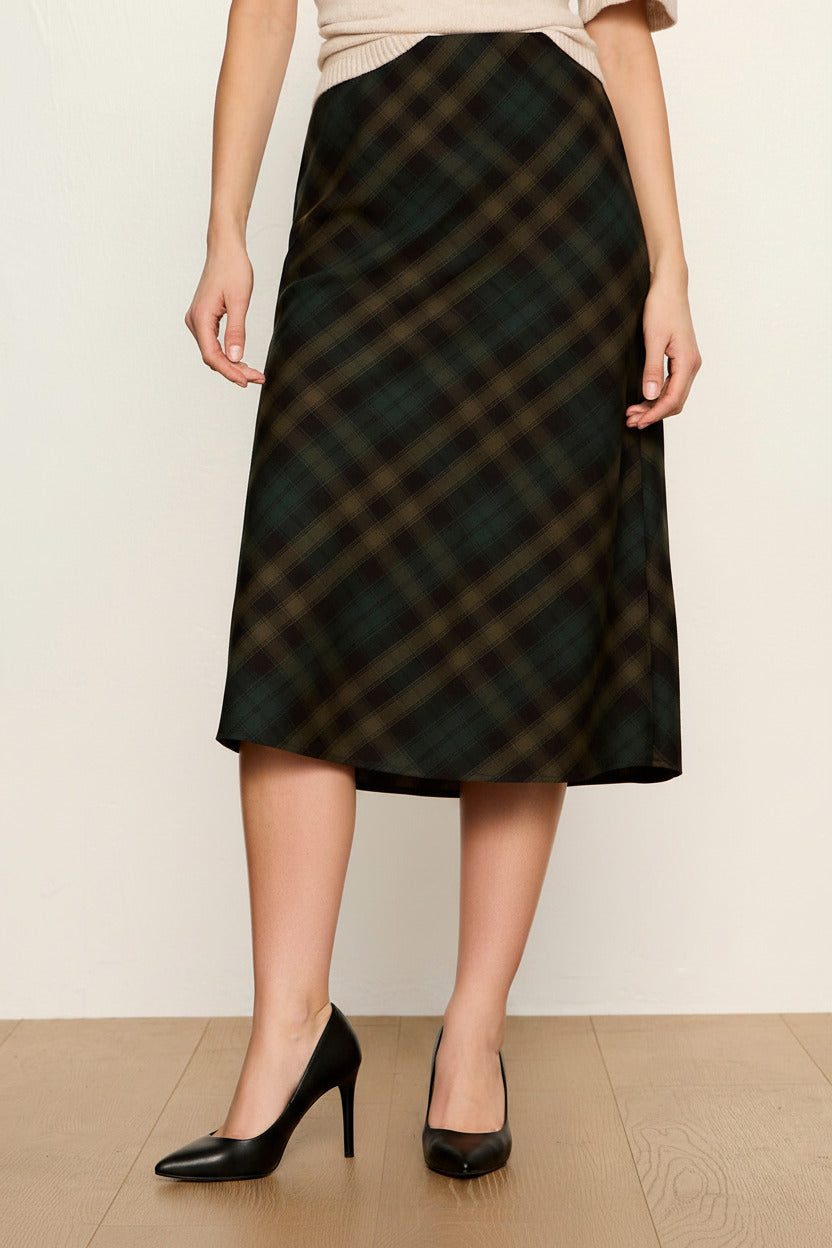 A person wearing a dark green and black plaid midi skirt with a beige top, black socks, and beige sneakers, standing against a plain off-white wall and light wood floor.