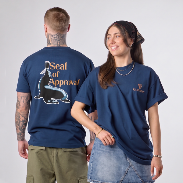 Two models wearing a navy blue t-shirt with a seal graphic on the back and a small crest logo on the front chest. The shirt features the text 'Seal of Approval'.
