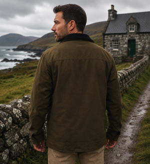 A man wearing an olive green corduroy jacket stands on a stone path in a coastal village, looking out towards the sea and a stone cottage.