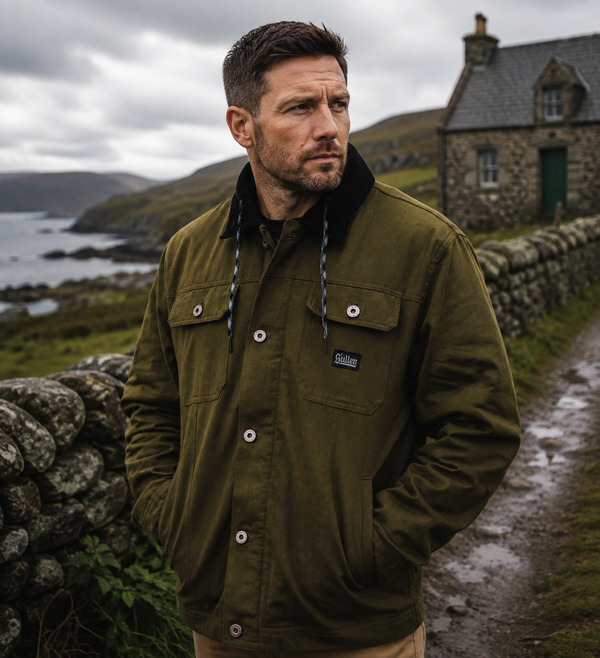 A man wearing an olive green Barbour overshirt jacket with a black fleece collar, standing outdoors on a stone path with a stone wall, sea, and cottage in the background.