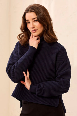 A woman wearing a dark navy ribbed knit mock neck sweater, posing with her hand near her chin against a light beige background.