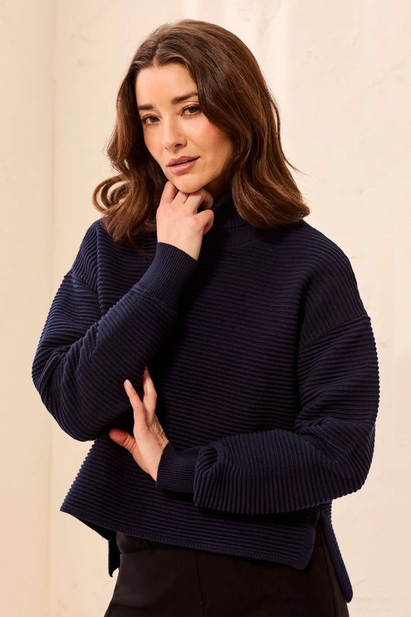A woman wearing a dark navy ribbed knit mock neck sweater, posing with her hand near her chin against a light beige background.