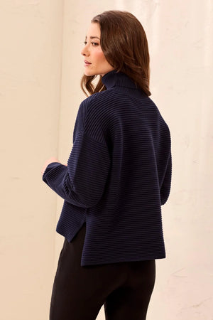 A woman wearing a navy blue ribbed knit mock neck sweater, shown from a side-back angle against a light beige textured wall.
