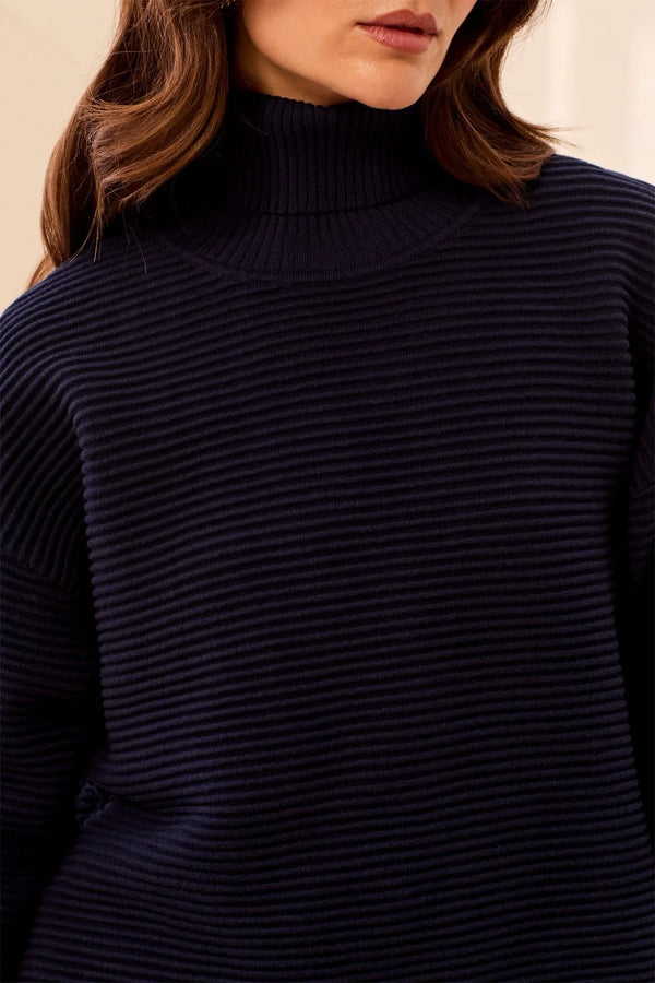 Close-up of a woman wearing a dark navy ribbed turtleneck sweater, showing the texture of the knit fabric and her hair against a soft neutral background.