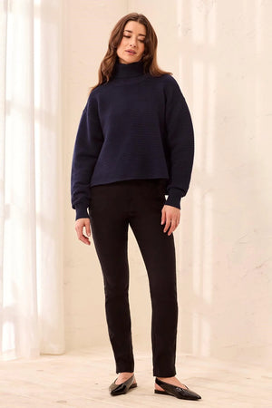 A woman modeling a dark navy ribbed knit mock neck sweater with black pants and black flats, standing in a brightly lit room with a textured cream wall.