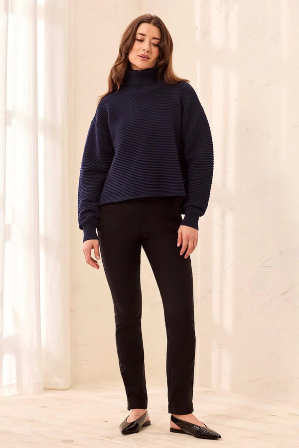 A woman modeling a dark navy ribbed knit mock neck sweater with black pants and black flats, standing in a brightly lit room with a textured cream wall.